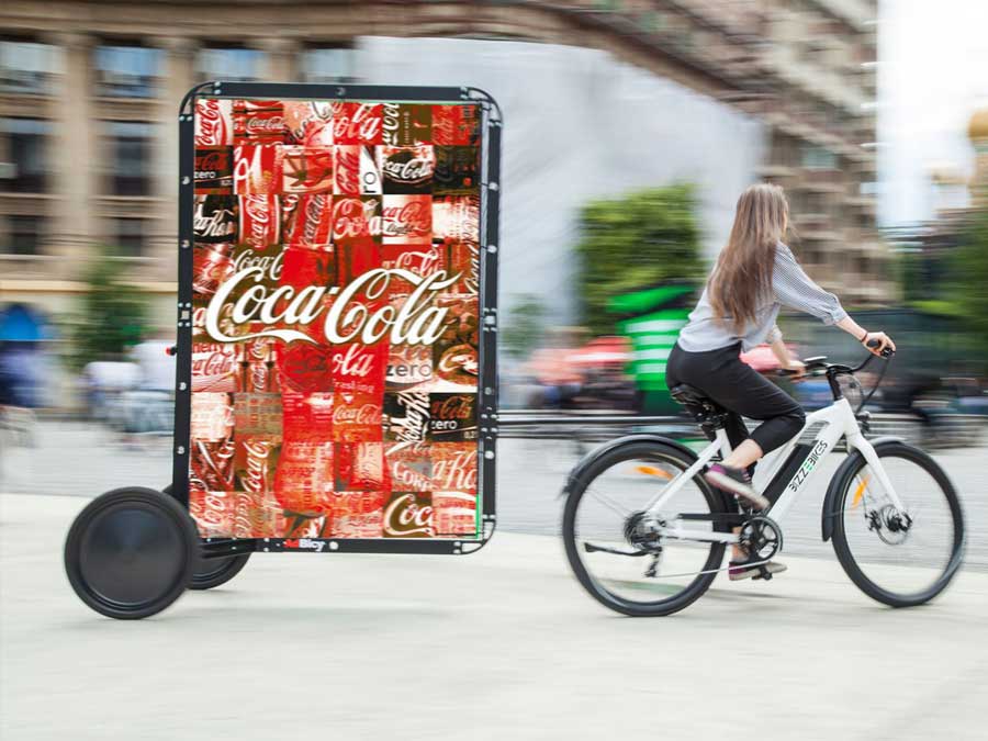 Chica montando su bicicleta adjunta a la publicidad de la calle Coca Cola.